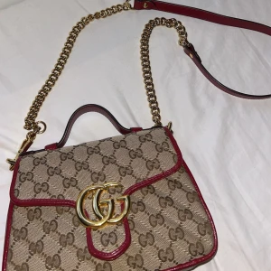 Gucci GG Supreme axelväska med kedja - Snygg axelväska från Gucci i klassiskt GG Supreme-mönster med beige och bruna toner, röda skinnkanter och guldfärgad kedja. Väskan har ett stort GG-spänne framtill och justerbar axelrem i både kedja och läder. Har tyvärr inte kvar box eller kvitto men qr kod finns inne i väskan. Kan skicka bild. Jätte fint skick nästan som ny. Köpt för 14000