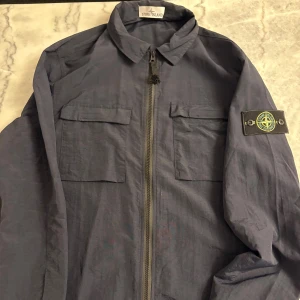 Blå overshirt från Stone Island - Snygg marinblå overshirt från Stone Island med klassisk krage, två bröstfickor med lock och dragkedja framtill. Ikonisk logotyp på vänster ärm. Perfekt för lager-på-lager och streetwear-stil.