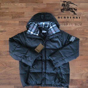 Burberry Jacka - Svart dunjacka från Burberry med klassisk Säljer en elegant och mångsidig Burberry-jacka i svart, i nyskick. Dunfodrad för värme och komfort med det klassiska rutmönstret i huvan som ger en exklusiv touch. Praktiska dragkedjefickor, avtagbar huva och diskret logopatch på ärmen. Sitter som (M/L). Jackan är i princip som ny och utan defekter. En lyxig kombination av stil och funktion – perfekt för både vardag och tillfällen då du vill klä upp dig.  📦Snabb leverans - skickas samma dag📦 