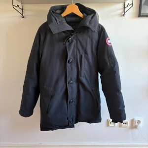 Svart dunjacka från Canada Goose - Säljer en svart dunjacka från Canada Goose med huva och klassisk logga på . flera fickor och en varm, quiltad insida. den har en lagning på vänster axel som är omärkbar. över de så har inte jackan några skador elle täcken på använding