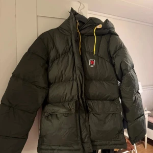 Grön Fjällräven  dunjacka XS - Säljer en grön Fjällräven Expedition Series dunjacka i storlek XS. Jackan har huva med gula snören, två stora fickor med lock och klassisk Fjällräven-logga på bröstet. Puffad design och dragkedja framtill. Perfekt för kalla dagar.