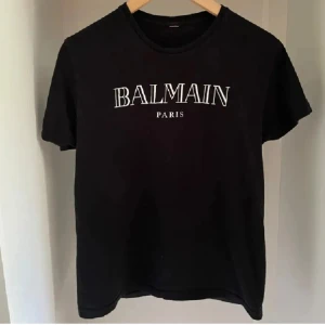 Svart Balmain Paris t-shirt - Snygg svart t-shirt från Balmain Paris med vit logga tryckt på bröstet. Klassisk rund halsringning och korta ärmar. Perfekt för dig som vill ha en stilren och trendig look med exklusiv känsla.