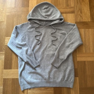 Grå kashmir hoodie - En enkel och stilren grå kashmir hoodie med huva och snörning framtill. Tröjan har långa ärmar och breda ribbade muddar vid ärmslut och nederkant. Perfekt för chill dagar och passar till det mesta. Materialet känns mjukt och är väldigt skönt enligt mig.