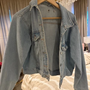 Ljusblå jeansjacka med knappar - Säljer en klassisk ljusblå jeansjacka med snygga silverfärgade knappar framtill och på fickorna. Jackan har en rak passform, krage och långa ärmar. Perfekt att slänga över en hoodie eller t-shirt för en chill look.