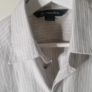 Randig kortärmad skjorta Haglöfs - Snygg kortärmad skjorta från Haglöfs i ljusgrått och vitt med tunna ränder. Skjortan har klassisk krage, knappar framtill och är tillverkad i ett lätt och luftigt material, perfekt för varma dagar.