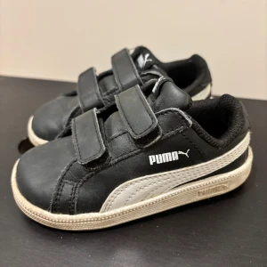 Svarta sneakers från Puma med kardborre - Svarta sneakers från Puma med vit sula och vit sidodetalj. Skorna har två praktiska kardborreband och är tillverkade i skinnliknande material. Klassisk Puma-logga på sidan och bekväm vadderad insida. Perfekta för en sportig och avslappnad stil.