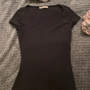 Svart ribbad topp från Anna Field - Svart ribbad topp från Anna Field med korta ärmar och v-ringad skärning. Toppen har en figurnära passform och är tillverkad i ett stretchigt material som sitter snyggt på kroppen. Perfekt att styla med jeans eller kjol för en trendig look.