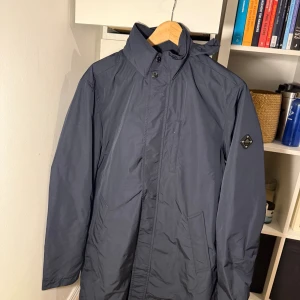 Mörkblå vindjacka från Peak Performance - Snygg mörkblå vindjacka från J Lindeberg med hög krage, diskret logga på ärmen och praktiska fickor. Jackan har dragkedja och knappar framtill samt en minimalistisk design som passar perfekt till vardagslooken.