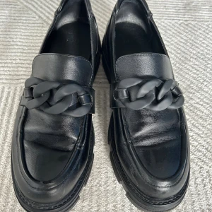 Chunky svarta loafers med kedjedetalj - Säljer ett par chunky svarta loafers i skinnimitation med grov sula och en stor kedjedetalj framtill. Skorna har rund tå och platt sula, vilket ger en cool och trendig look. Perfekta för dig som vill sticka ut med dina skor.