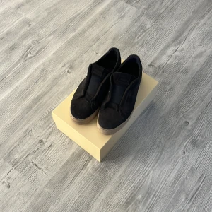 Svarta Axel Arigato sneakers med platåsula - Svarta slip-on sneakers från Axel Arigato med platåsula i beige. Ovandel i mocka och insida i skinn, minimalistisk design utan snörning. Perfekta för dig som gillar stilrena och trendiga skor med lite extra höjd.