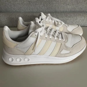 Adidas Cloudfoam vita sneakers - Snygga Adidas Cloudfoam sneakers i vitt och beige med klassiska tre ränder på sidan. Ovandel i mesh och mocka för en fräsch look och extra komfort. Platt sula med Cloudfoam-dämpning och snörning framtill. Perfekta för en clean och sportig stil. Storlek 40 2/3. 