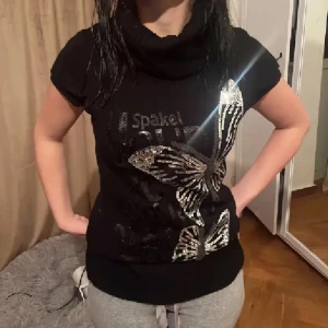 Svart topp/klänning med paljettfjärilar - Säljer en svart topp/klänning med korta ärmar och stor polokrage. Toppen har snygga paljettfjärilar i silver framtill och textdetaljer. Modellen är längre och sitter tight mot kroppen. Perfekt för dig som gillar lite extra bling i vardagen. Kan användas som topp eller klänning. Vintage❤️
