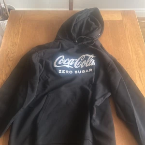 Svart Coca-Cola Zero Sugar hoodie - Svart hoodie med stor Coca-Cola Zero Sugar-logga i vitt framtill. Klassisk huva med snörning och känguruficka. Perfekt för dig som gillar streetwear och vill sticka ut med en ikonisk logga. Mjuk och skön, passar till chill dagar.