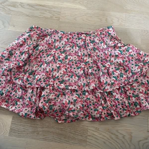 Blommig volangkjol från Zara - Supersöt kort volangkjol från Zara med blommigt mönster i rosa, rött, grönt och vitt. Kjolen har flera lager volanger som ger en flowy känsla och är perfekt för varma dagar. Tillverkad i ett lätt och luftigt material.