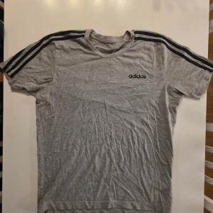 Grå Adidas t-shirt med svarta ränder - Snygg grå t-shirt från Adidas med klassiska svarta ränder längs axlarna och svart Adidas-logga på bröstet. T-shirten har rund halsringning och är kortärmad. Perfekt för en sportig och avslappnad stil.                                                                                     STORLEK M