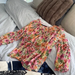 Blommig blus från Zara med volang - Färgglad blus från Zara med blommigt mönster i rosa, orange, grön och gul. Långa, luftiga ärmar och volangdetalj vid halsen samt knyt framtill. Tunt, lätt material som ger en flowy känsla. Perfekt för dig som gillar romantiska och färgstarka plagg. Guldiga detaljer är också med i sömmarna. PRIS GÅR ATT DISKUTERA