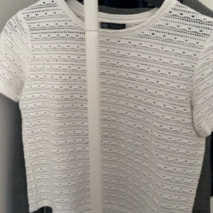 Vit spetsig topp från Zara - Säljer en vit topp från Zara med korta ärmar och rund hals. Toppen har ett snyggt hålmönster i vågiga rader som ger en fräsch och luftig känsla. Perfekt att styla med jeans eller kjol för en trendig look.