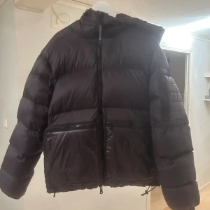 Svart pufferjacka från J.Lindeberg - Säljer en svart pufferjacka från J.Lindeberg med huva och stora framfickor med dragkedja. Jackan är fodrad med ett mönstrat innerfoder i rött och olivgrönt/svart. Bara änvänd några få gånger. Perfekt för kalla dagar och riktigt snygg street/stockholmsstil. Kvitto finns och pris kan diskuteras vid snabb affär! 