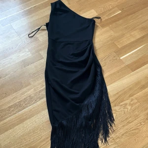 Svart oneshoulder klänning med fransar - Snygg svart oneshoulder klänning med asymmetrisk skärning och långa fransar längs nederkanten. Klänningen har en smal axelrem på ena sidan och är figurnära med draperad detalj på höften. Perfekt för dig som vill sticka ut med en cool och trendig look.