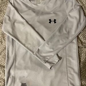 Ljusgrå långärmad träningströja UA - Säljer en ljusgrå långärmad träningströja från Under Armour. Tröjan har rund halsringning, svart logga på bröstet och är tillverkad i ett lätt och ventilerande syntetmaterial som passar perfekt för träning. Snygg och enkel design med diskreta sömmar.