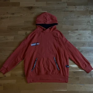 Rostfärgad hoodie från WOXO 720° - Säljer en snygg rostfärgad hoodie från WOXO 720° med svart innerfoder i huvan och coola detaljer med text på fickorna. Tröjan har känguruficka, tryck på bröstet och ribbade muddar. Perfekt för dig som gillar streetstyle och vill ha något bekvämt och trendigt.