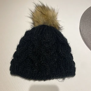 Svart stickad mössa med fuskpälsboll - Mysig svart stickad mössa med grovt kabelstickat mönster och en stor, fluffig fuskpälsboll i beige/brun nyans på toppen. Insidan är fodrad med mjuk, svart fleece för extra värme. Perfekt för kalla vinterdagar och snygg till alla ytterkläder.
