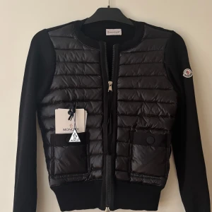 Svart cardigan från Moncler - Svart cardigan från Moncler med quiltad framsida i blankt material och stickade ärmar. Koftan har två fickor framtill med knapp, dragkedja och Moncler-logga på ärmen. Storlek 1, använd endast 1 gång då den var för liten för mig, så precis som ny! 🩷