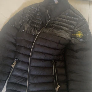 Svart dunjacka från Stone Island - Säljer en svart dunjacka från Stone Island med klassisk patch på ärmen. Jackan har glansiga partier upptill, dragkedja framtill och två fickor med dragkedja. Perfekt för kalla dagar och har en riktigt clean look.
