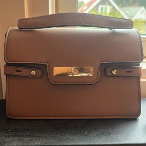 Brun handväska från Primark - Snygg brun handväska från Primark med stilren rektangulär form och guldfärgade metalldetaljer. Väskan har ett praktiskt handtag upptill och dekorativa remmar framtill. Perfekt för dig som gillar klassisk och enkel design.