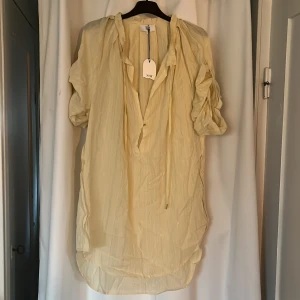 Ljusgul glittrig kortklänning från NOA NOA - Superfin ljusgul klänning från NOA NOA med glittriga ränder i tyget. Klänningen har v-ringning med knytband, korta ärmar med knappdetalj och en avslappnad passform. Perfekt för sommarens alla tillfällen!