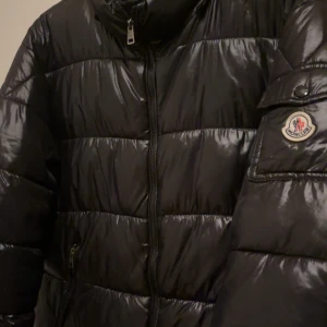 Svart pufferjacka från Moncler - Säljer en svart, glansig pufferjacka från Moncler med klassisk logga på ärmen. Jackan har dragkedja framtill, hög krage och två fickor med dragkedja. Perfekt för kalla dagar och riktigt snygg streetstil.