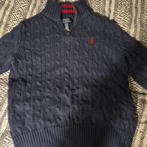 Mörkblå stickad tröja Polo Ralph Lauren - Kabelstickad mörkblå tröja från Polo Ralph Lauren med halv dragkedja och hög krage. Klassisk röd logga broderad på bröstet och ribbade muddar. Perfekt för kyliga dagar och snygg till jeans.