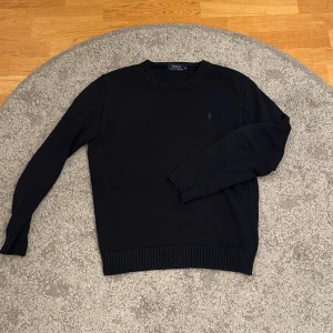  tröja från Polo Ralph Lauren - Marinblå stickad tröja från Polo Ralph Lauren med rund hals och diskret grön logobrodyr på bröstet. Tröjan har ribbade muddar vid ärmslut och nederkant, vilket ger en snygg och klassisk look. Perfekt för dig som gillar stilrena plagg.