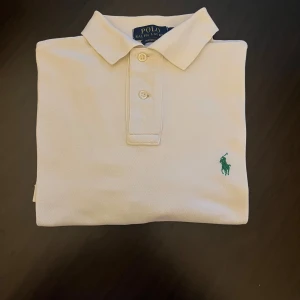 Vit pikétröja från Polo Ralph Lauren - Klassisk vit pikétröja från Polo Ralph Lauren i bomull med grön broderad logga på bröstet. Tröjan har krage, knappar framtill och custom fit-passform. Perfekt för en clean och stilren look.