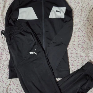 Svart träningsset från Puma - Säljer ett svart träningsset från Puma med vit logga på både jacka och byxor. Jackan har dragkedja och vita paneler upptill, byxorna har resår i midjan och dragsko. Perfekt för dig som gillar sportig stil och vill ha något bekvämt och snyggt.