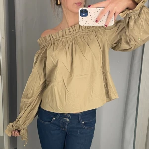 Beige offshoulder blus med puffärm - Säljer en beige blus med offshoulder och bred resår runt halsen. Blusen har långa puffärmar med snörning vid ärmslutet som ger en cool och trendig look. Materialet är lätt och känns skönt mot huden, perfekt för dig som gillar avslappnad stil.
