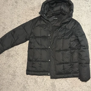 Svart pufferjacka från Lyle & Scott - Svart pufferjacka från Lyle & Scott med huva och quiltad design. Jackan har långa ärmar och tryckknappar framtill. På ärmen finns den klassiska gula Lyle & Scott-loggan. Perfekt för kalla dagar och har en clean, modern look.