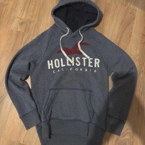 Vintage Hollister hoodie - Snygg vintage hoodie från Hollister med stor logga och röd fågel på bröstet. Tröjan har huva med vita snören, magficka och mjukt fleecefoder på insidan. Perfekt för chill dagar och extra mysig tack vare det fluffiga materialet.