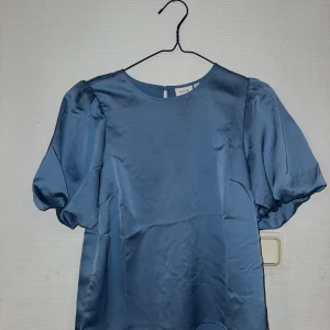 Blå satinblus med puffärm från Vila - Säljer en stilren blå blus från Vila i glansigt satinmaterial. Blusen har rund halsringning, korta puffärmar och en enkel knapp i nacken. Perfekt för dig som gillar en clean look med lite extra volym på ärmarna. Använd en gång! 