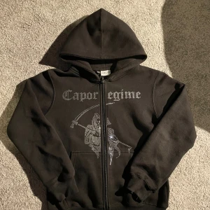 Svart hoodie Capor Regime Costino XS - Svart hoodie från Costino med glittrande Capor Regime-tryck och motiv av en lieman i strass på framsidan. Tröjan har huva, dragkedja och långa ärmar. Perfekt för dig som gillar edgy streetstyle och vill sticka ut med coola detaljer.