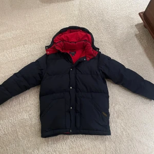 Blå pufferjacka från Polo Ralph Lauren - Säljer en mörkblå pufferjacka från Polo Ralph Lauren med röd insida och huva. Jackan har långa ärmar, två stora fickor framtill och stängs med både dragkedja och tryckknappar. Perfekt för kalla dagar och har en schysst, klassisk look. Storlek large i barnstolek så xs-s i vanlig storlek.  Tveka inte på o skriva vid minsta fundering