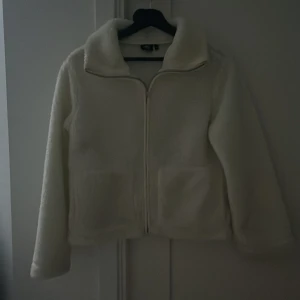 Vit teddyjacka från 157, XS - Mysig vit teddyjacka från 157 i storlek XS. Jackan har en rak passform, två stora fickor framtill och dragkedja hela vägen upp. Perfekt för dig som gillar fluffiga och stilrena jackor med krage.