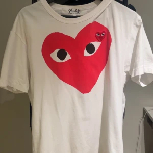 Cdg t shirt  - Säljer en vit t-shirt från Comme des Garçons Play med det ikoniska röda hjärtat med ögon tryckt på bröstet och ett mindre broderat hjärta ovanför. T-shirten har rund hals och korta ärmar, tillverkad i mjuk bomull. 