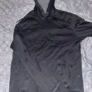 Svart hoodie från Hugo Boss - Svart hoodie från Hugo Boss med broderad logga på bröstet. Tröjan har huva med snörning och en stor magficka framtill. Insidan är grå och materialet känns mjukt och bekvämt, perfekt för chill dagar. Stilren design som funkar till jeans eller joggers.