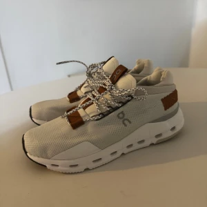 On Cloudnova sneakers i beige och brunt - Säljer ett par On Cloudnova sneakers i beige mesh med bruna detaljer och vit chunky sula. Skorna har svartvitmönstrade snören och coola detaljer på hälen. Perfekta för dig som gillar sportig och modern stil.