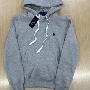 Grå hoodie från Polo Ralph Lauren - Snygg grå hoodie från Polo Ralph Lauren med klassisk broderad logga på bröstet. Tröjan har huva med vita snören, känguruficka och ribbade muddar. Perfekt för en chill och stilren look.