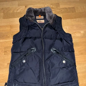 Fräsch och ny äkta Parajumper väst. - Svart dunväst från Parajumpers i slim fit med mjuk fuskpäls på kragen och dragkedja framtill. Västen har två snedställda fickor med dragkedja och tryckknapp, samt detaljer i orange. Perfekt för kalla dagar när du vill ha en sportig och snygg look. #Äkta