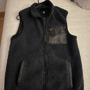 Svart fleeceväst med dragkedja - Säljer en svart fleeceväst med dragkedja och praktiska fickor. Västen har en bröstficka med dragkedja och är perfekt för kyligare dagar. Köparen står för frakt!