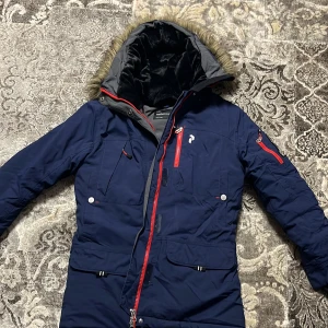 Mörkblå Peak Performance dunjacka S Woman - Peak Performance dunjacka i mörkblått med röda dragkedjor och flera fickor. Jackan har huva med fuskpäls, quiltad insida och lång ärm. Perfekt för kalla dagar med snygg design och praktiska detaljer. Kan prata om priset.