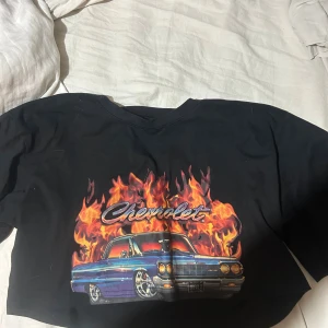Svart croppad Chevrolet t-shirt - Svart croppad t-shirt med fet Chevrolet-print framtill, där en blå bil och eldflammor ger en riktigt cool vibe. Kortärmad och tillverkad i mjuk bomull, perfekt för dig som gillar streetstyle och bilar. Snygg passform som sitter loose. Storleken är M eller L